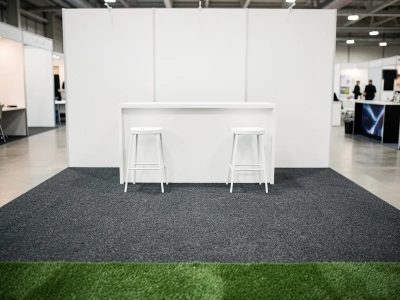Messestand med mørkegråt messetæppe og en stribe kunstgræs foran, hvid disk og to barstole i en messehal