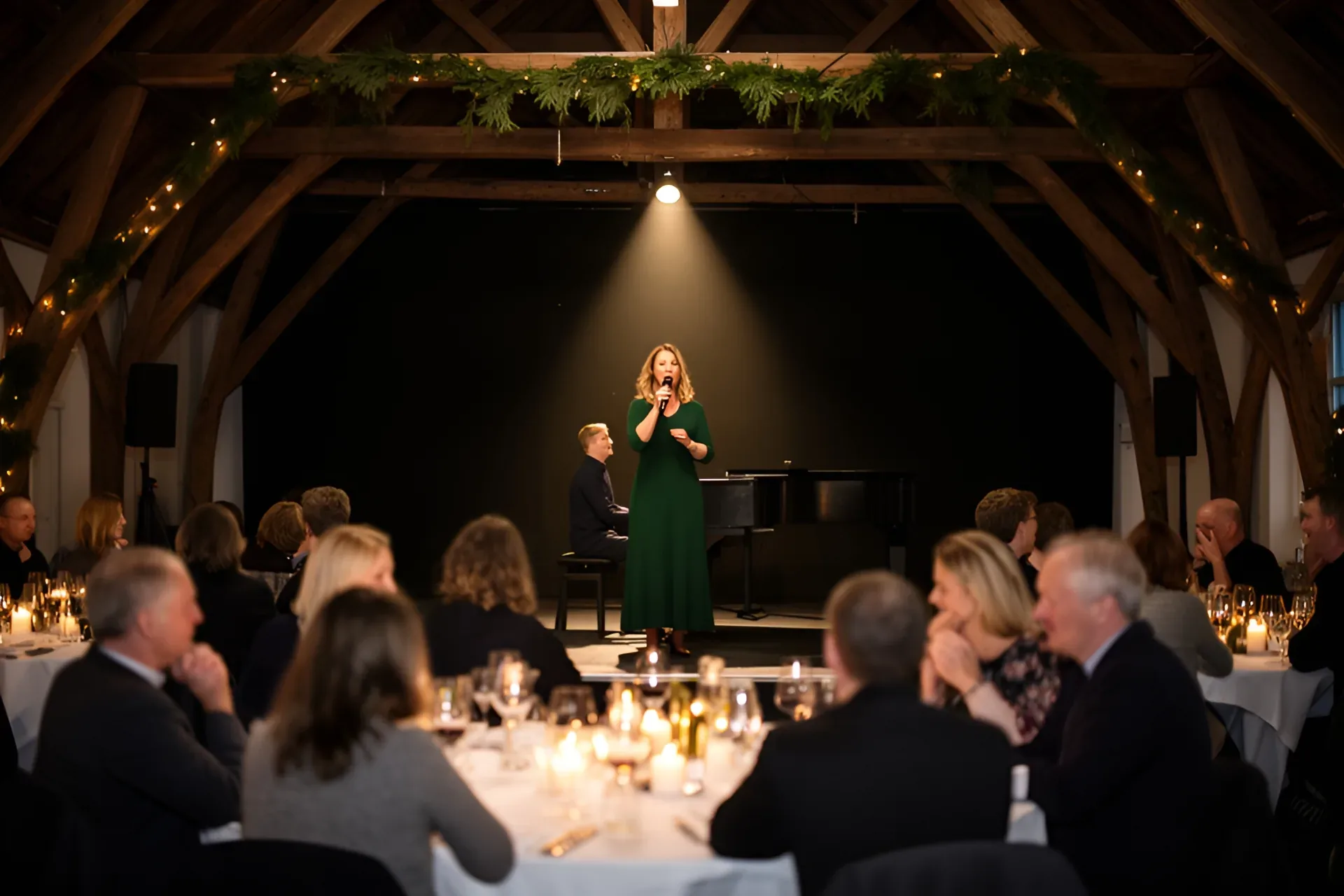 En sangerinde i grøn kjole synger i en håndmikrofon på en lav scene ved en firmajulefrokost i en sal med bjælker og granguirlander, en pianist spiller ved flyglet, og kollegerne lytter ved runde borde med hvide duge og levende lys