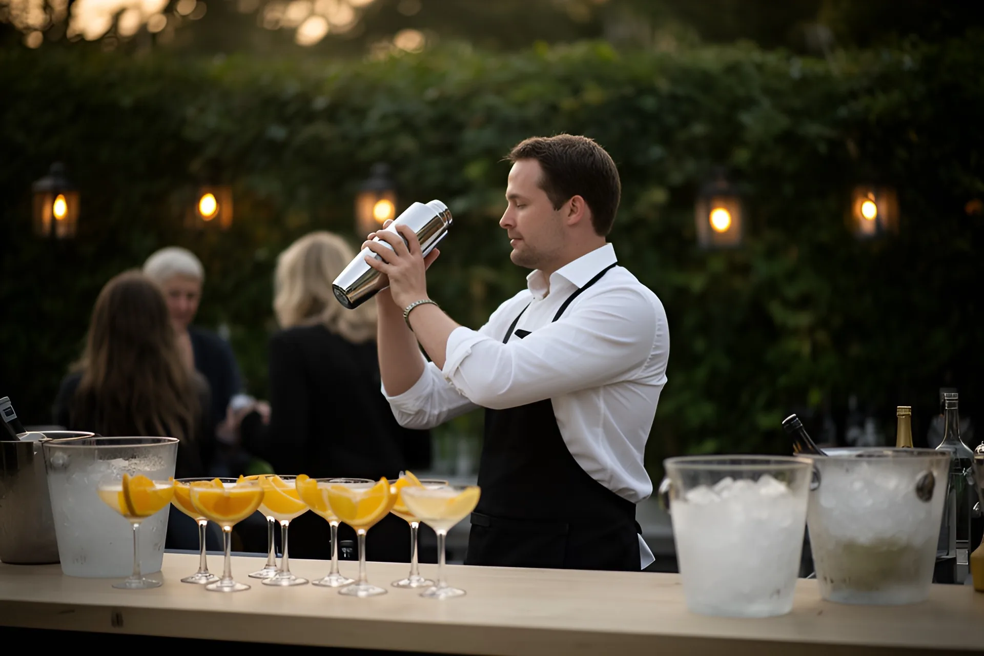 En bartender i hvid skjorte og sort forklæde ryster en cocktailshaker bag en lys træbar med en række coupeglas med appelsinskiver og to isspande, gæster og lanterner i den uskarpe hæk bagved