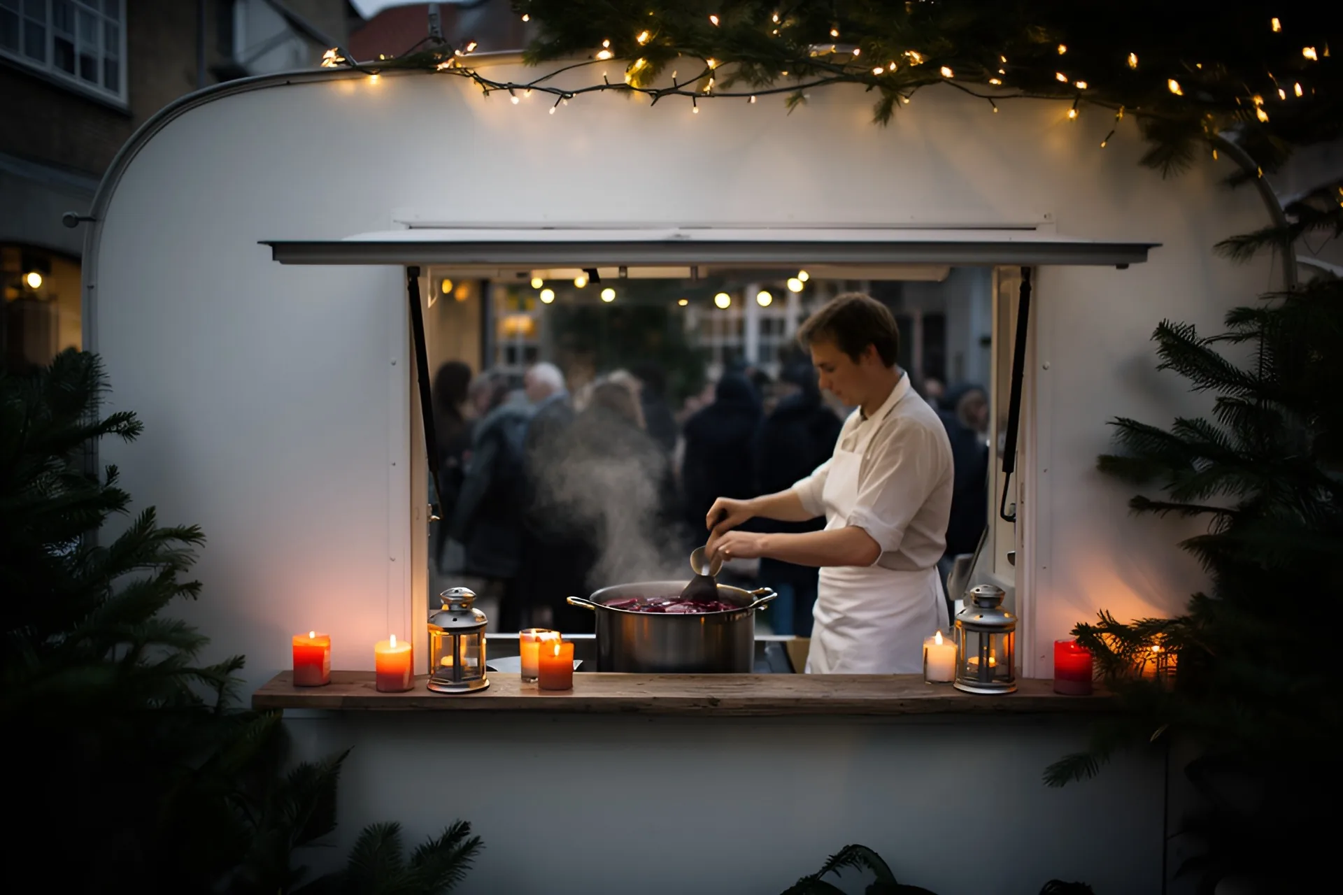 Hvid foodtruck-lem i en gård i december i skumringen, en kok i hvid kittel rører i en stor gryde med dampende gløgg, lanterner med stearinlys og grangrene langs disken og lyskæder over, gæster som sløret silhuetter i baggrunden