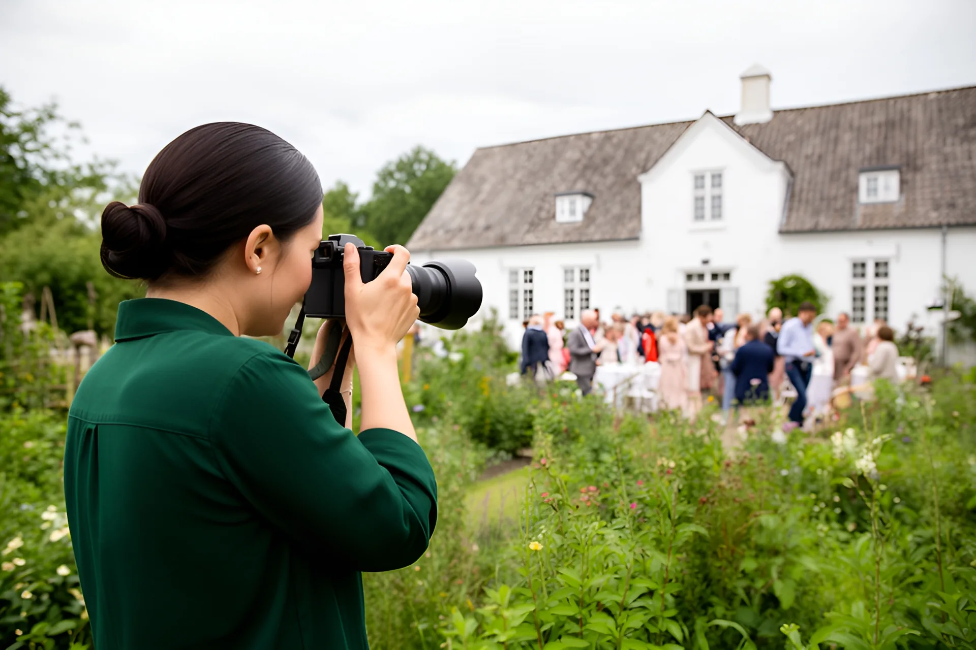 En fotograf i grøn skjorte, set bagfra, fotograferer et bryllupsselskab i haven ved et hvidkalket stuehus på en gråvejrsdag, gæsterne står ved de dækkede borde i baggrunden