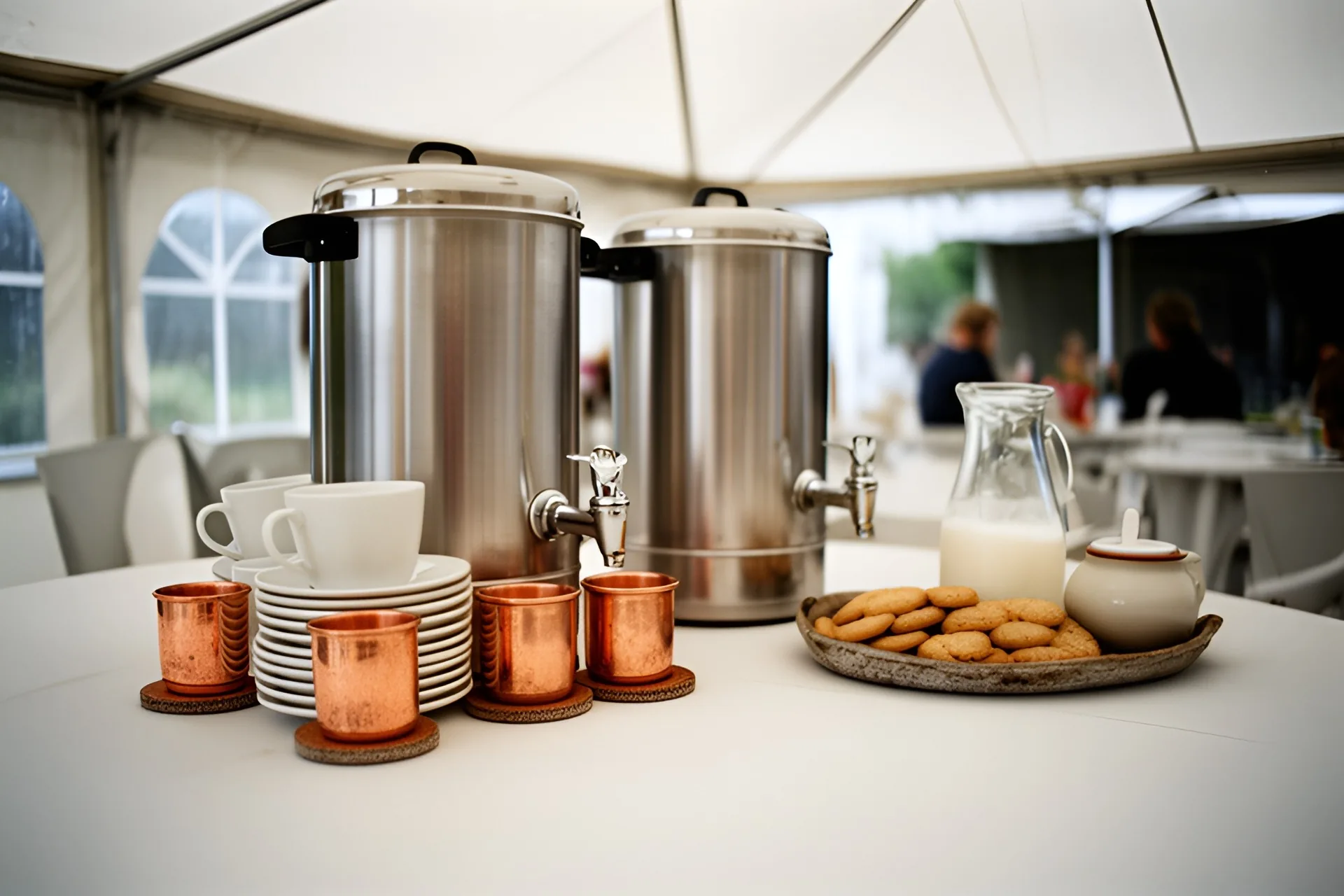 To store rustfri kaffe-termokander med haner, kopper på underkopper, kiks, mælkekande og sukkerskål på et bord i festtelt