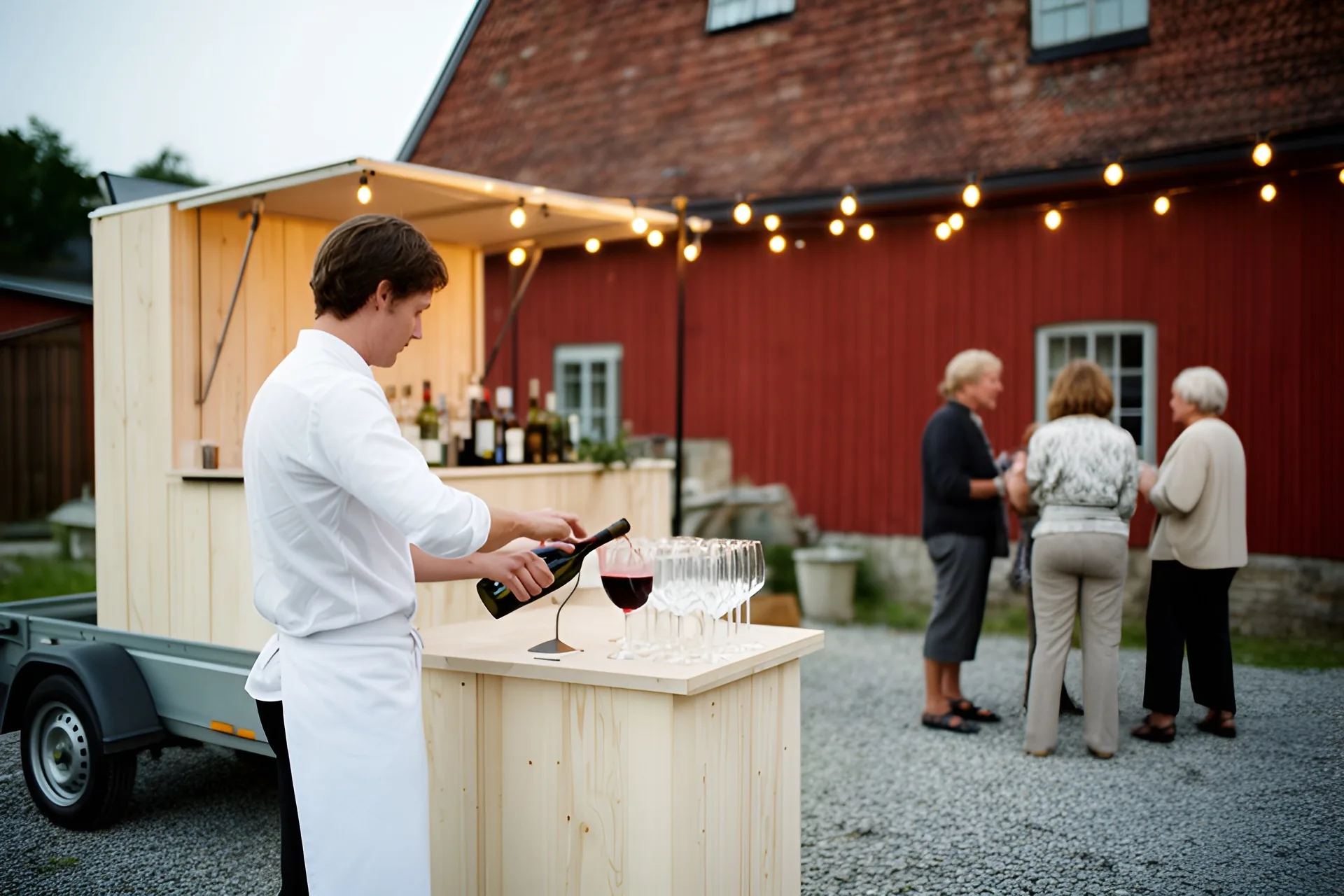 En bartender i hvid skjorte og forklæde hælder rødvin i et glas ved en mobil bar af lyst træ på en anhænger, en række vinglas på disken, tre gæster taler sammen på gruset foran en rød lade, lyskæder over baren