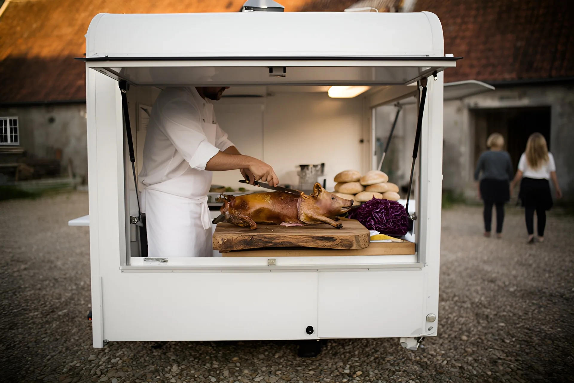 Hvid foodtruck med åben lem på en grusbelagt gårdsplads, en kok i hvid kittel skærer i en helstegt pattegris med sprød svær på et stort skærebræt, rundstykker og rødkål ved siden af, to gæster går bort set bagfra