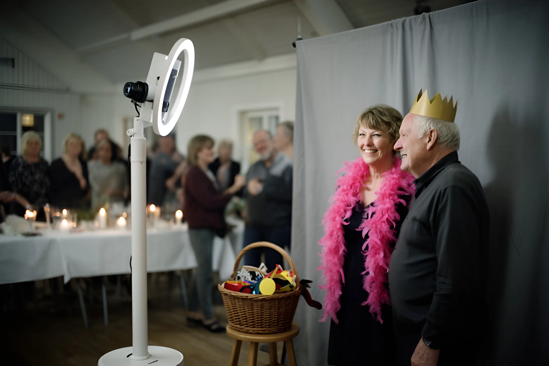 En photobooth med kamera og ringlys på en hvid stander i et lyst forsamlingshus, hvor en kvinde med lyserød fjerboa og en mand med en guldkrone af papir smiler foran en grå fotobaggrund, mens en kurv med rekvisitter står på en skammel og gæsterne ved festbordene ser til bagved