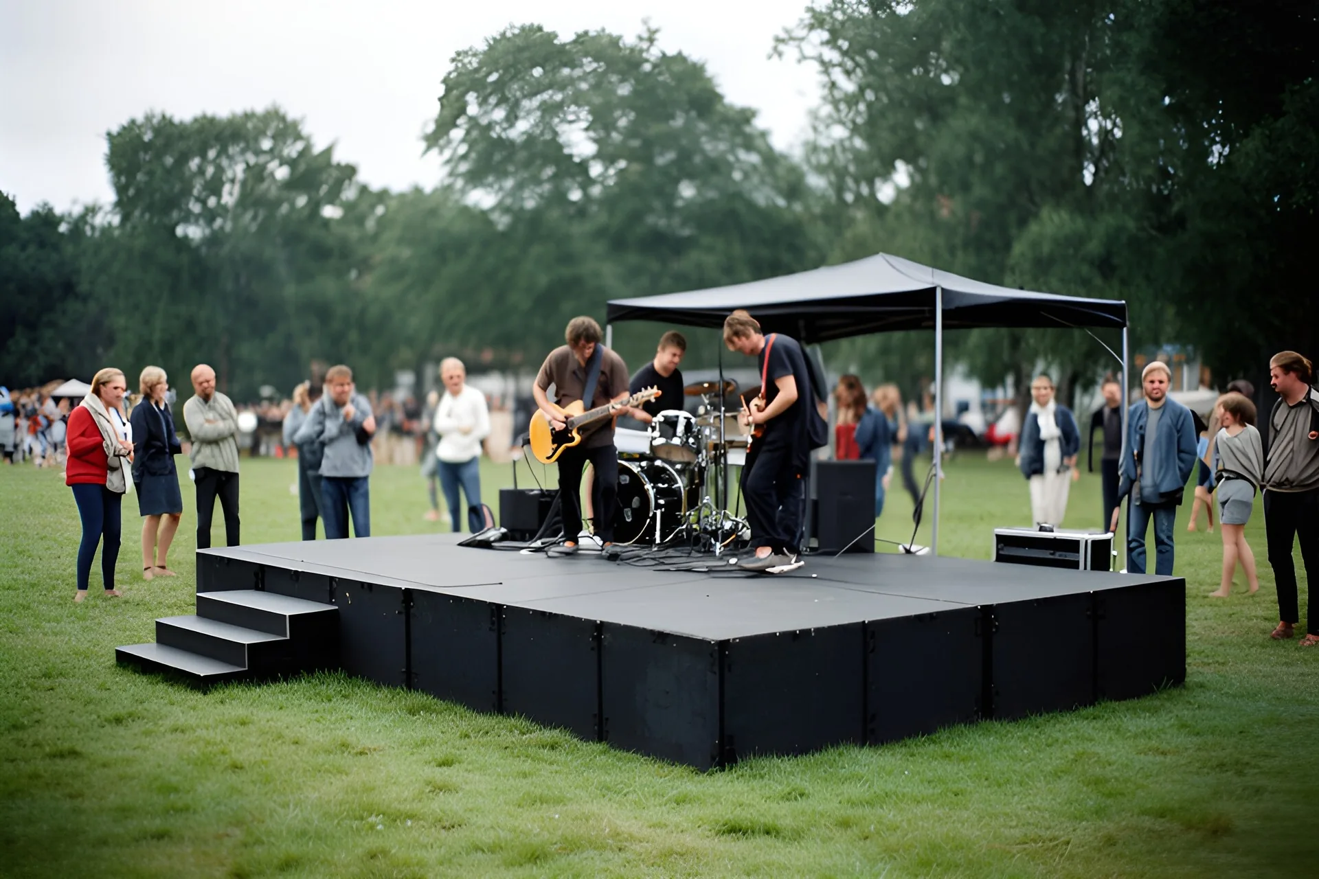 Lav sort scenepodie med trappe på en græsplæne og et lille sort halvtag, et band på tre stiller op, gæster står på afstand.