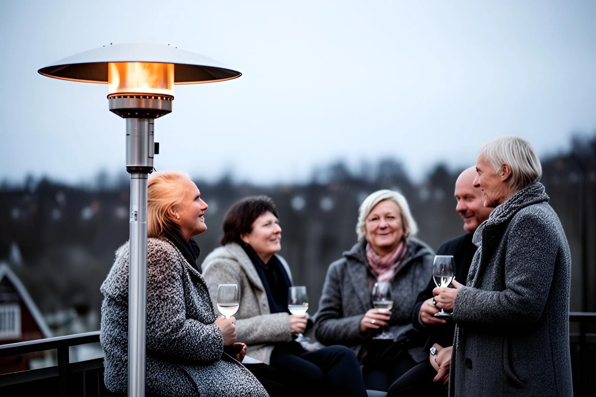 Gæster i uldjakker står og sidder og snakker med vinglas ved en høj terrassevarmer en kold aften, lyskæde i baggrunden.