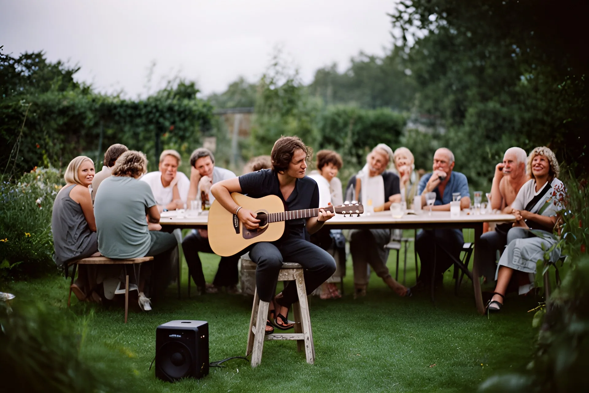 En ung mand spiller akustisk guitar på en skammel for gæster ved et langbord i en dansk have en sommeraften.