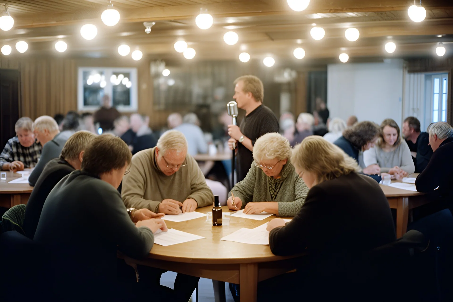 Quizaften i et dansk forsamlingshus, voksne sidder i grupper ved runde borde og skriver svar på papir, en quizmaster med en gammeldags mikrofon står for enden af salen under varme loftslamper
