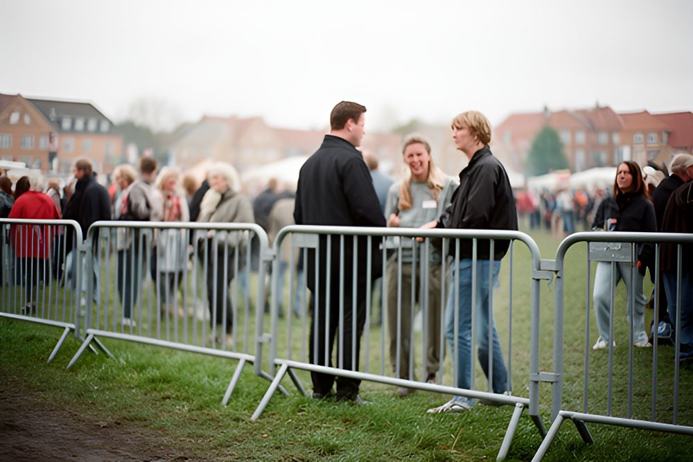 Sølvfarvede afspærringshegn på en græsplæne ved en byfest, gæster samlet bag hegnet