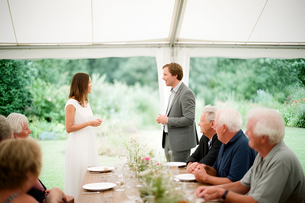 Gæster taler ved et langbord med vilde blomster i et hvidt telt med åbne sider i en have
