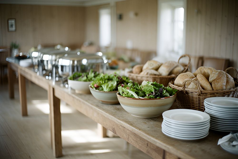 Buffetbord med salatskåle, brød i kurve, kantiner og stakke af hvide tallerkener i et lyst forsamlingshus