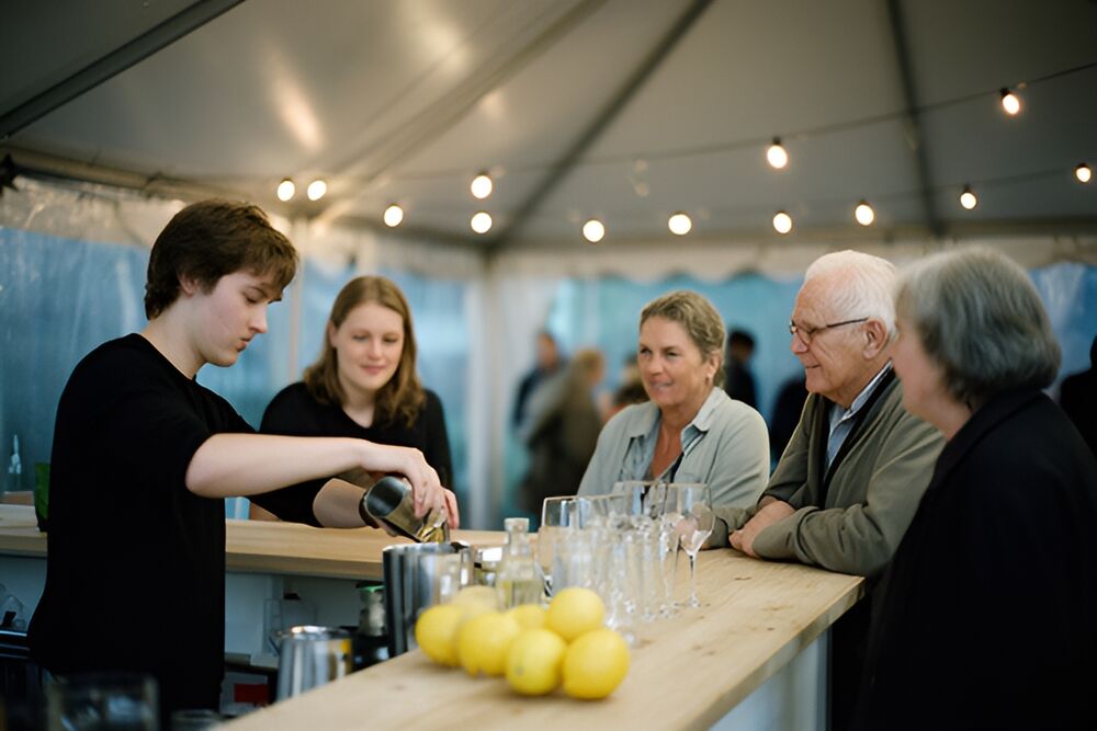 Bartender hælder en cocktail fra en shaker ved en lys træbardisk, mens gæster lytter og smiler