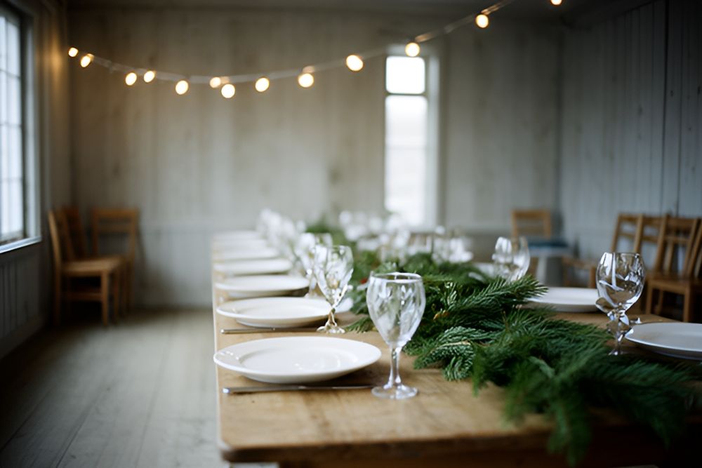 Langbord med hvide tallerkener, vandglas og grangrene i et lyst forsamlingshus, lyskæde under loftet
