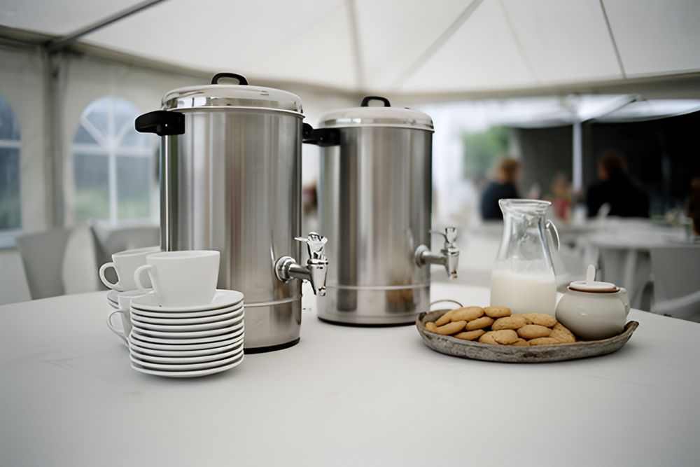 To store rustfri kaffe-termokander med haner, kopper på underkopper, kiks, mælkekande og sukkerskål på et bord i festtelt