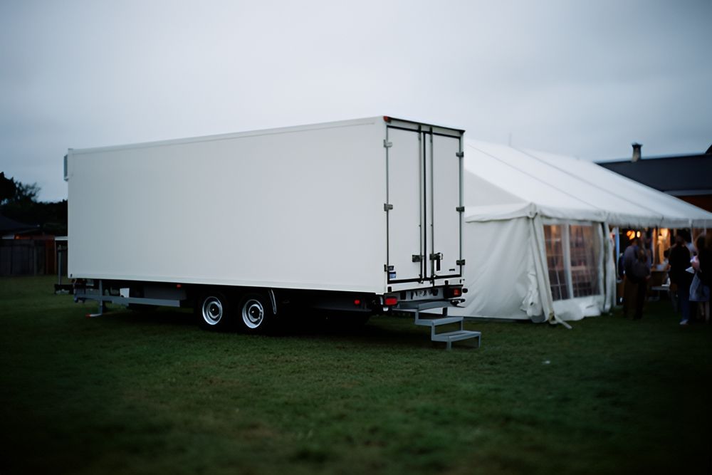 Hvid kølertrailer med bagdøre og trappetrin parkeret på en græsplæne ved siden af et hvidt festtelt en overskyet aften
