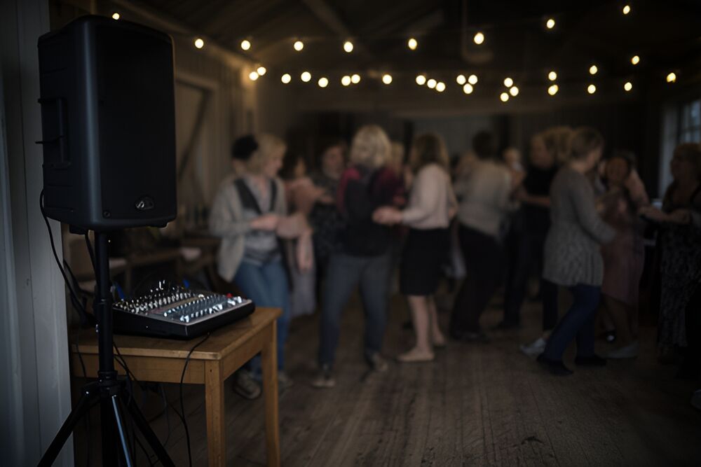 Sort aktiv højttaler på stativ og en lille mixer på et træbord i et forsamlingshus om aftenen, mens gæster danser i baggrunden under lyskæder.