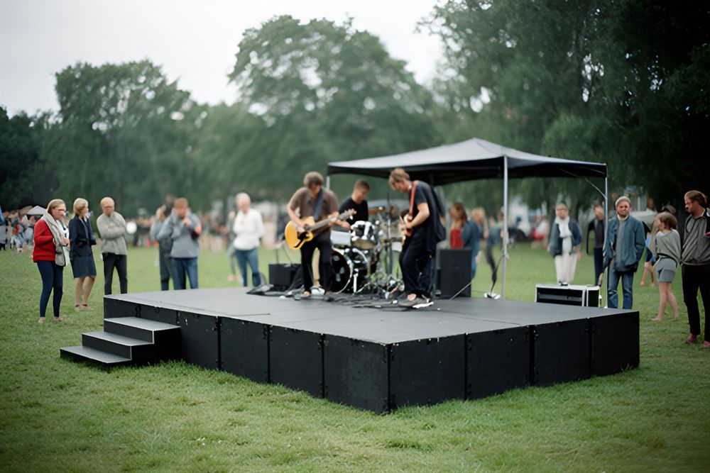 Lav sort scenepodie med trappe på en græsplæne og et lille sort halvtag, et band på tre stiller op, gæster står på afstand.