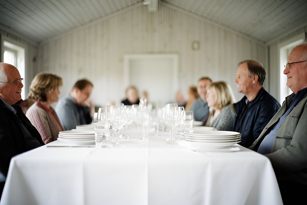 Langbord med hvid dug, tallerkener og glas i et forsamlingshus, gæster sidder ved bordet