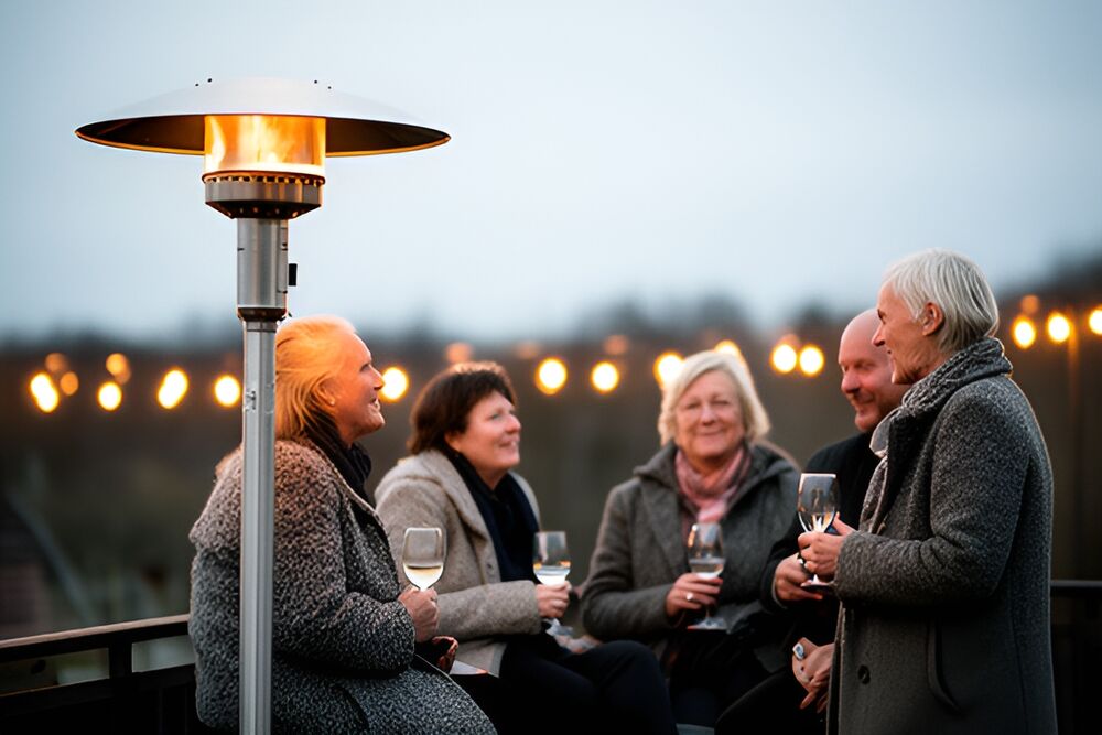 Gæster i uldjakker står og sidder og snakker med vinglas ved en høj terrassevarmer en kold aften, lyskæde i baggrunden.