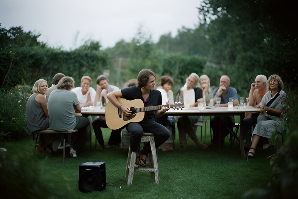 En ung mand spiller akustisk guitar for gæster ved et langbord i en have en sommeraften